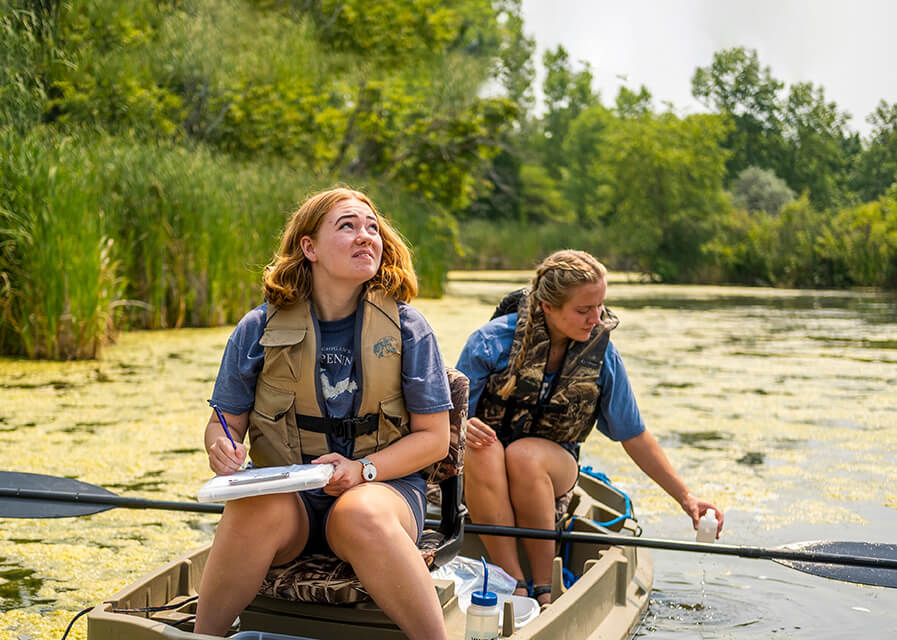 Two students canoe through a wetland area with one taking notes while the other collects a water sample.