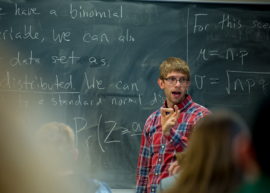 Mathematics faculty member standing in front of chalk board while speaking to a class.