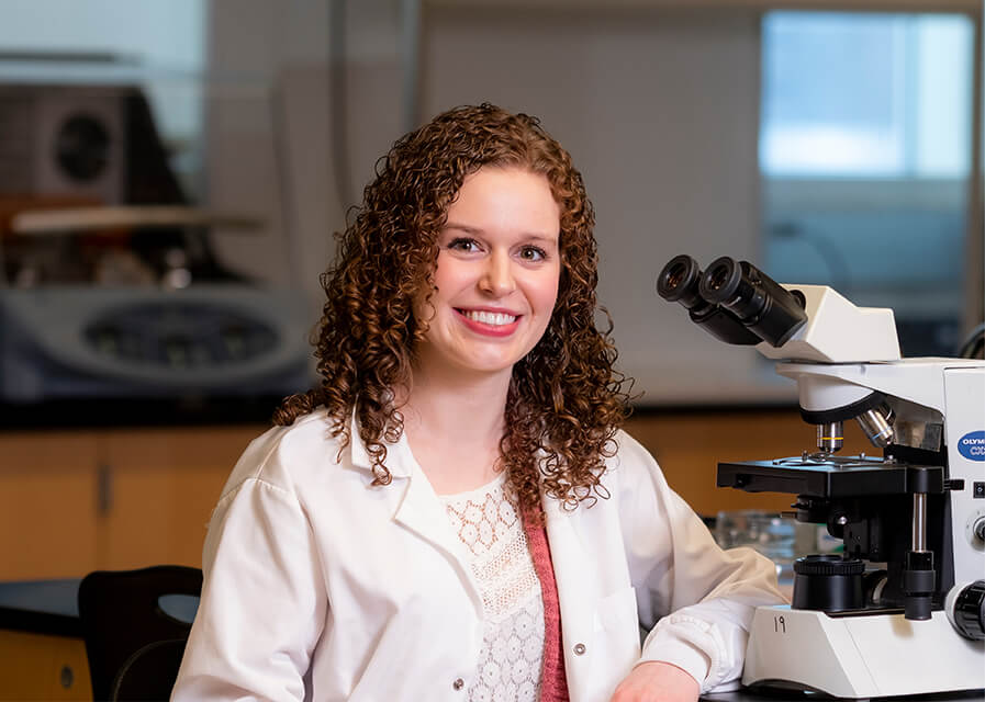 A pre-med student in a white lab coat sits beside a microscope.