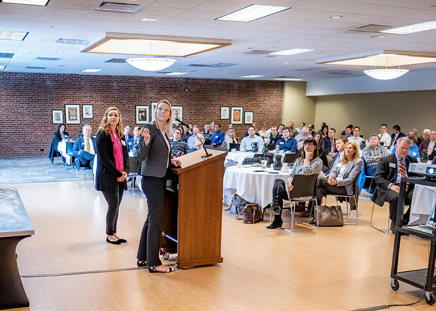 Two female students present to a room of area professionals.