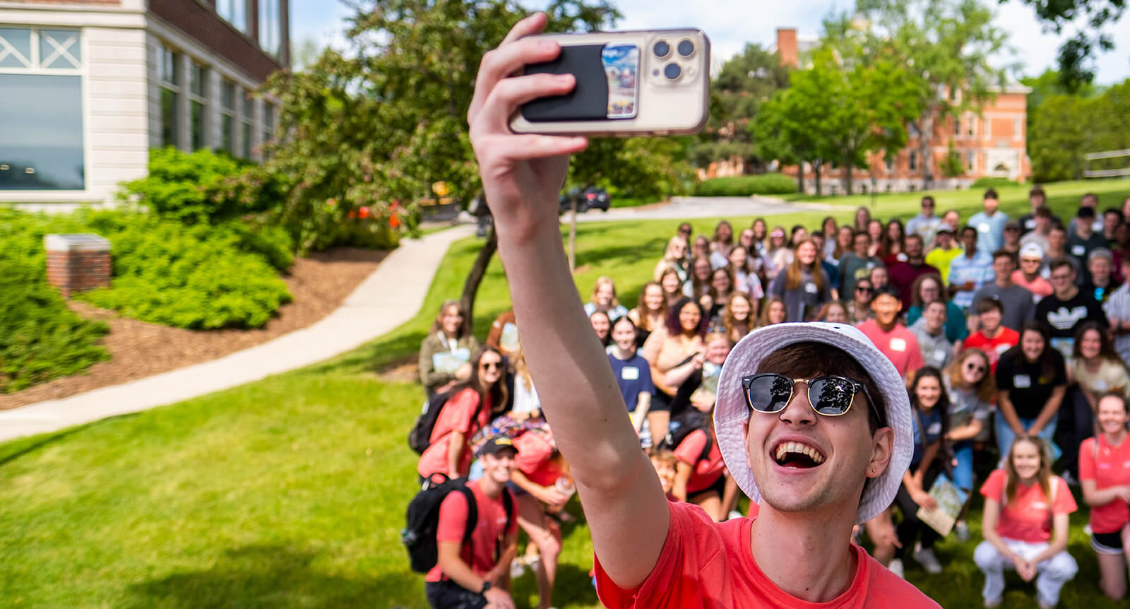 Student stands in front of a larger crowd of students holding up his phone to take a selfie.