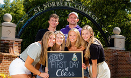 Students gather around a sign about the first day of classes to pose in front of the SNC arch
